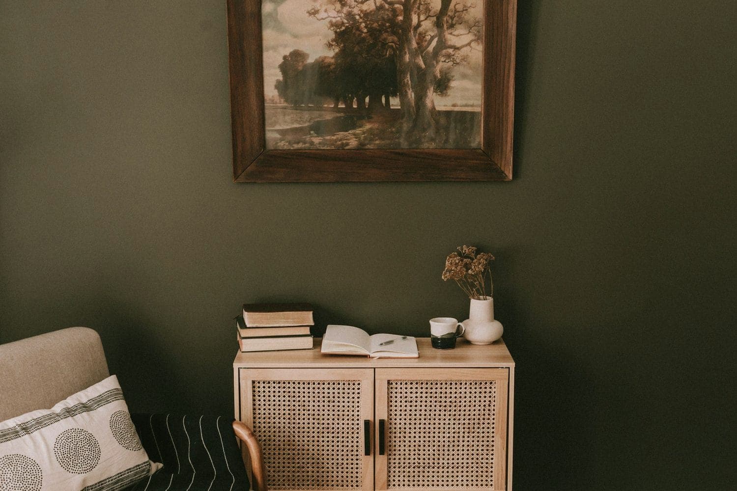 A dark green room with a framed landscape above a cane cabinet